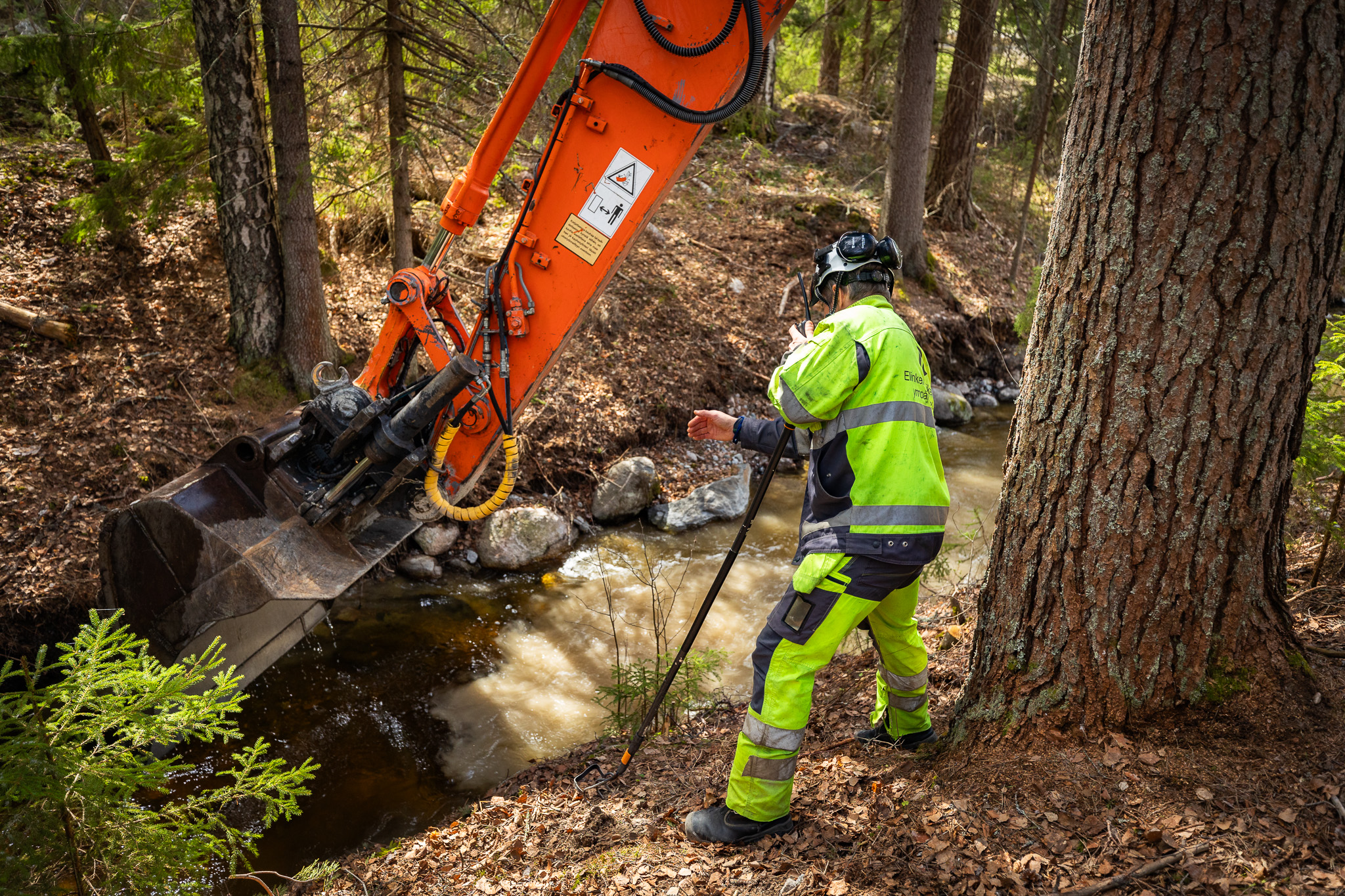 Kaivinkoneen kauha kaivaa puustoisessa ympäristössä ojaa, jossa on vettä. Työvaatteisiin pukeutunut henkilö seisoo ohjan reunalla nojaten työvälineeseen. Kuva vain luonto- ja ilmastoviestintään.