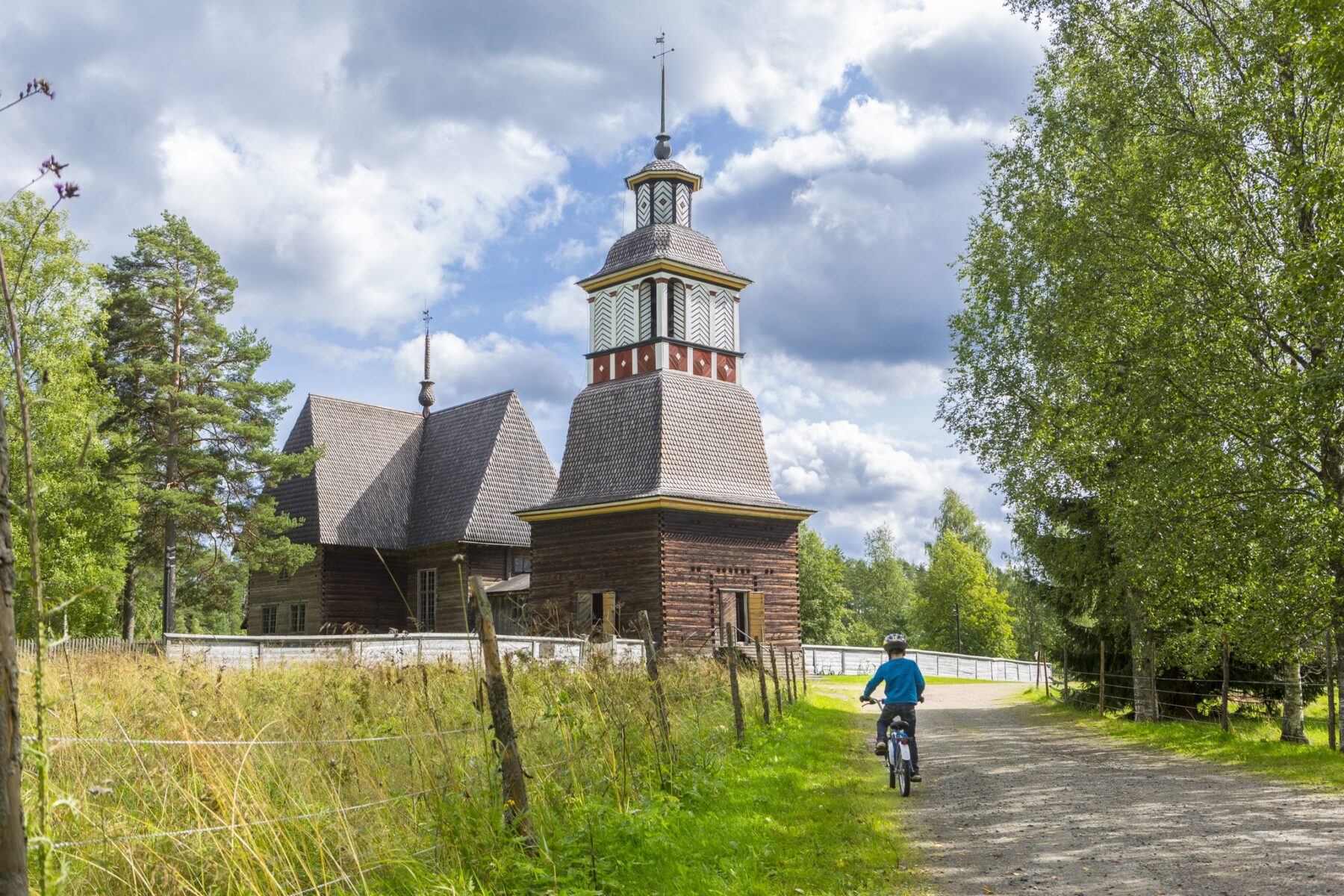Pellon reunalla vanha puukirkko. Vieressä kasvaa puita. Kirkolle vievällä hiekkatiellä pyöräilee lapsi.
