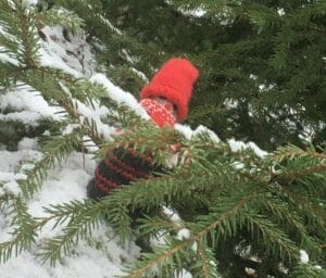 Käsitöin tehty tonttu lumisella maalla kuusen alla.